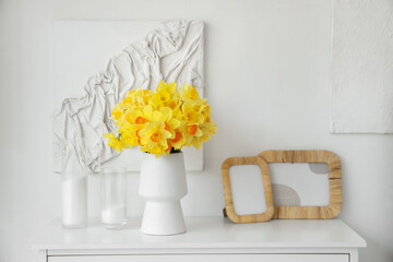 Vase with daffodils, candles and frames on commode in living room