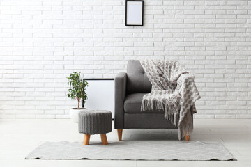 Armchair with plaid, pouf and houseplant with frame near white brick wall