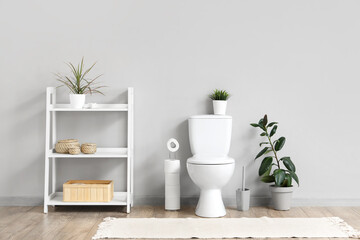 Interior of restroom with toilet bowl, shelving unit and plant near grey wall