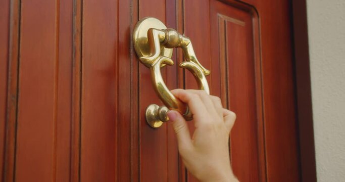 A hand holding a brass door knocker on a wooden door, preparing to knock. Close-up of aesthetic ancient architecture.