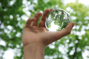 Beautiful green trees outdoors, overturned reflection. Man holding crystal ball in park, closeup