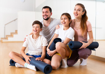 Smiling fit woman and man with teenage girl and boy posing with yoga mats before training in modern...