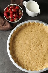 Making shortcrust pastry. Raw dough in baking dish, milk and strawberries on grey table, top view