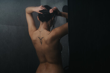 An artistic shot capturing the rear view of a woman while showering, focused on the water droplets on her skin in a shadowy, intimate setting.
