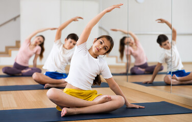 Smiling teen girl doing Padmasana with side stretch during yoga workout in gym with family..