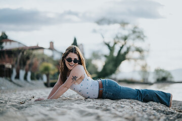 A cheerful young woman lies on a sandy beach under a clear sky, wearing sunglasses and a smile,...