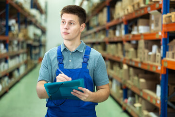 In warehouse rack area of paint store, guy worker in blue overall checks availability and quantity...