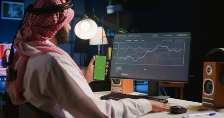 Man holding green screen smartphone while looking at business annual report graphs on computer....
