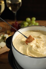 Dipping piece of bread into fondue pot with melted cheese at table, closeup