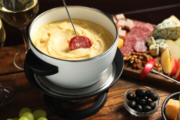 Fork with piece of sausage, melted cheese in fondue pot and other products on wooden table, closeup
