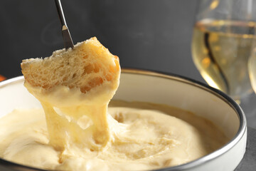 Dipping piece of bread into fondue pot with melted cheese on grey background, closeup