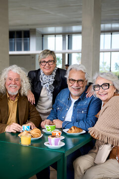 Vertical. Portrait group seniors Caucasian people posing together sitting at cafeteria. Older friends with gray hair looking smiling at camera with breakfast on table. Happy matures in nursing home