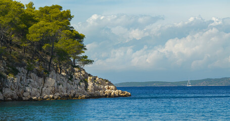 Pine trees on a rocky island coast and a sailboat sailing on the blue open sea. A beautiful sunny and windy day for exploring and cruising around the picturesque Dalmatian islands in Adriatic Sea.