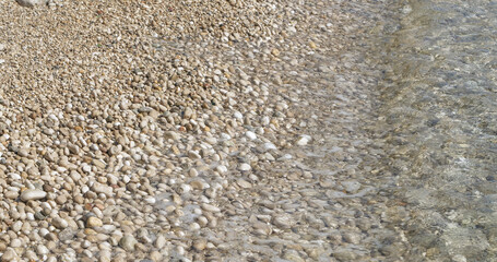 CLOSE UP: A peaceful shore where soft waves roll over a natural mosaic of sunlit pebbles, creating a harmonious and textured coastline scene perfect for relaxation. Meditative feel from the seaside.