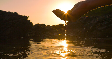 CLOSE UP, LENS FLARE: Sunbeams peep through a hand scooping water in sea puddle formed at low tide...