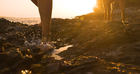 LENS FLARE, CLOSE UP: Barefoot woman walks with her dog on a rocky beach at sunset. Rugged Adriatic...