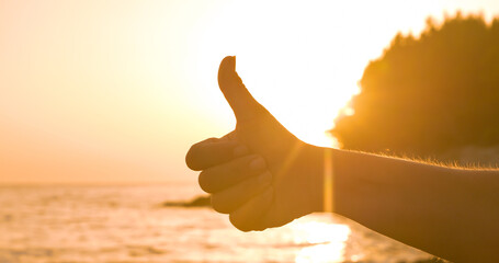 LENS FLARE, SILHOUETTE: A thumbs up against the tranquil sunset by the ocean. Familiar hand gesture...