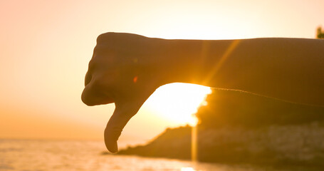 LENS FLARE, SILHOUETTE: Golden lit thumbs down gesture on the beach at sunset. Close up view of a...