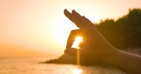 LENS FLARE, CLOSE UP, SILHOUETTE: Hand gesture precisely frames the setting sun against seascape...