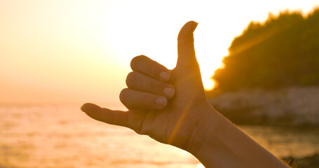 LENS FLARE, CLOSE UP, SILHOUETTE: Morning sunrise on beach and shaka hand gesture expressing good...