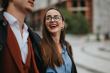 Cheerful woman with glasses smiling and talking with a man outdoors in an urban environment,...