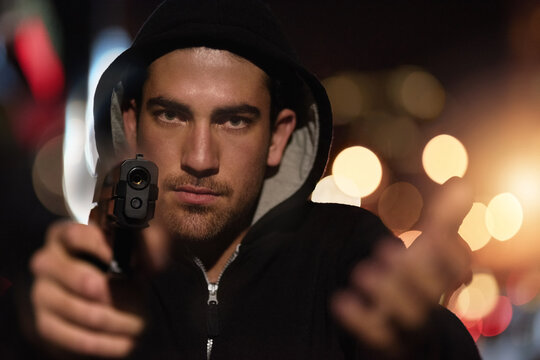 Man, portrait and gun point in city as gangster thief with bokeh lights at night or robbery pistol, hoodie or dark. Male person, face and hand or downtown for dangerous crime or steal, thug or theft