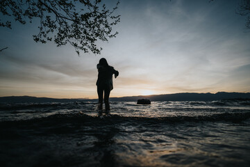 Fototapeta premium A young girl stands by a lake at sunset, splashing water gently, surrounded by nature, conveying joy and tranquility.