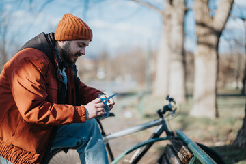 Focused man with beard in a warm orange beanie and red jacket using a smart phone, sitting by his...