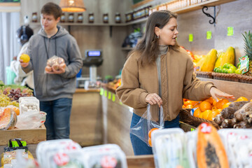 Careful young female buyer purchasing oranges in grocery store with large assortment