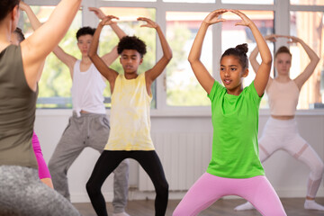 Teenage African American girl in vibrant workout clothes practicing basic ballet moves with other beginners at group class led by female choreographer..