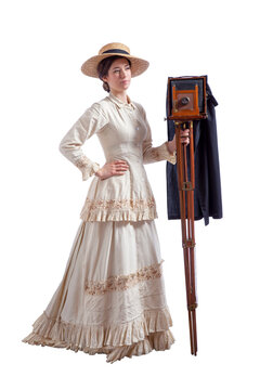 Young woman with camera wearing a beige vintage 1880s dress and straw hat isolated on white