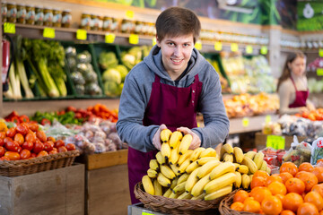 Young salesman in apron lays out bananas on counter in vegetable shop..