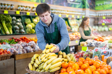 Positive young salesman placing bananas in a market basket in large grocery store