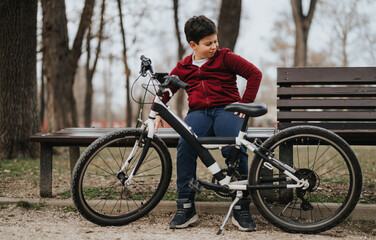 Fototapeta premium A young boy takes a break on a park bench beside his bicycle, enjoying a moment of leisure outdoors.