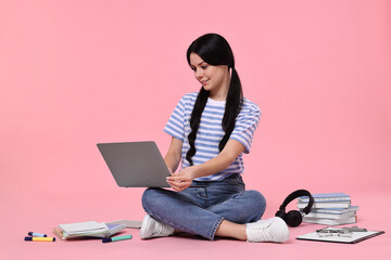 Student with laptop sitting among books and stationery on pink background