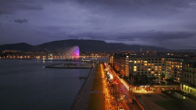 Stunning aerial view of Geneva's lit waterfront at twilight, showcasing vibrant streets, buildings, and the iconic jet d'eau fountain