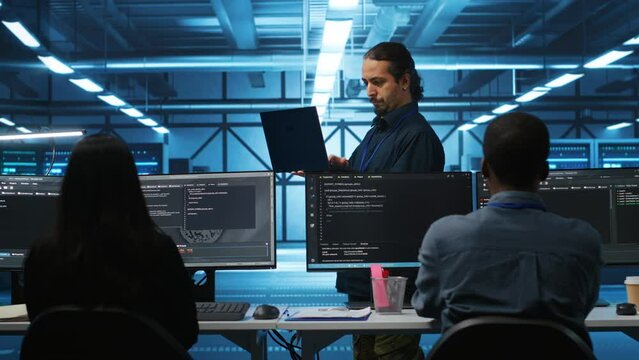 Middle eastern manager in server hub workplace doing inspection on electronics and multiracial workers. Team leader reading paperwork on laptop, verifying infrastructure documents, doing audit