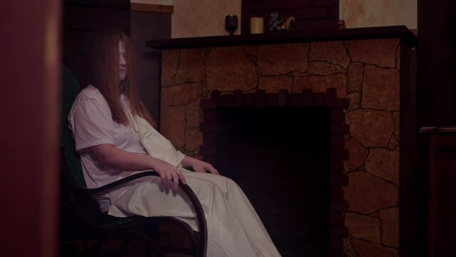 Creepy woman with pale face dressed in white nightgown slowly swings on rocking chair. Performance of professional actress in quest room