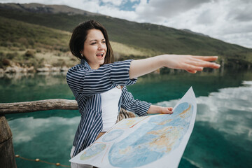 An enthusiastic woman traveling stands by a lake, holding a map and pointing to the destination,...