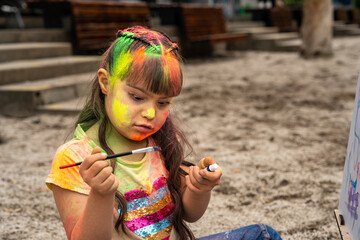Beautiful little girl with Down syndrome painting on canvas outdoors in a sandbox in a public park...