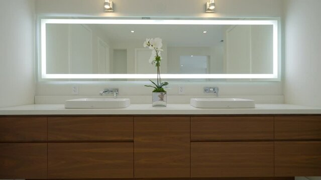 Modern bathroom vanity with large mirror framed by bright led light. Two white sinks with silver faucets and wooden cabinets with several drawers