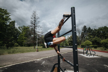 Muscular man trains outdoors, executing an inverted maneuver on fitness equipment in a lush green...