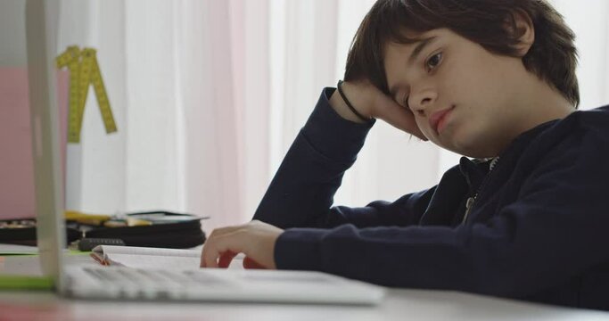 A young boy appears bored as he rests his head on his hand, working on homework with his laptop in a bright room.