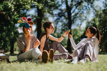 Three young friends sit on the grass in a sunny park, sharing a joyful moment while playing with...