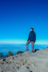 an Asian man taking a photo at the top of Mount Ciremai wearing a navy jacket and cream cargo pants with a beautiful view and a very beautiful clear blue sky