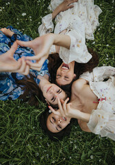 Fototapeta premium A heartwarming moment captured as three young girls lie on the grass, laughing and making peace signs, surrounded by small flowers in a park.