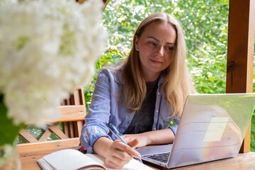Young happy woman focuses on her laptop in wooden alcove. Relaxed outdoor setting emphasizes...