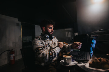 A concentrated male professional works on a laptop in a dimly lit room, with headphones around his neck and a cup of coffee nearby. © qunica.com