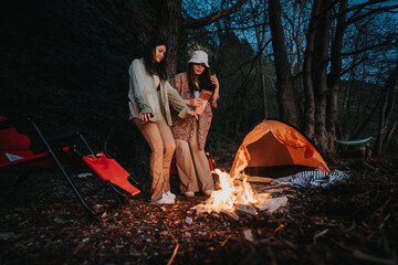In this image, two women enjoy the tranquil ambiance of a forest camping scene by a gently...