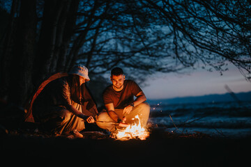 Two friends preparing dinner over a campfire by a lake, enjoying the peaceful outdoors at night.